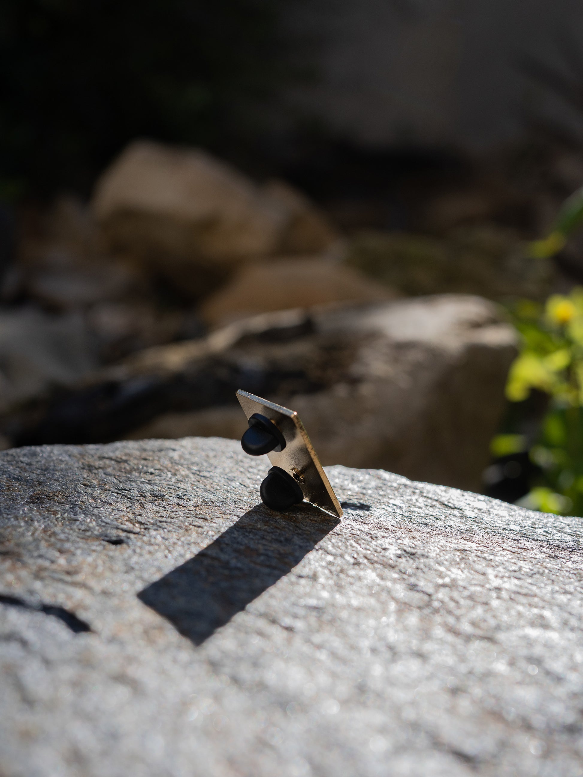 Hitorii "Napping Gou" metal chalkbag pin shown from the back on a backdrop of a rock in sunlight. The pin has a plain metal back with two pins, lined up vertically, for extra stability.