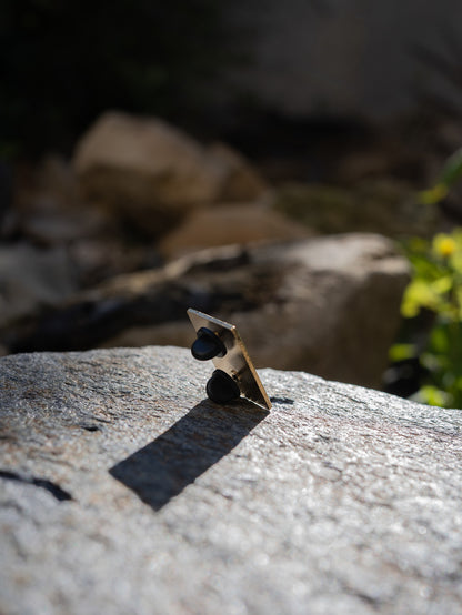 Hitorii "Napping Gou" metal chalkbag pin shown from the back on a backdrop of a rock in sunlight. The pin has a plain metal back with two pins, lined up vertically, for extra stability.